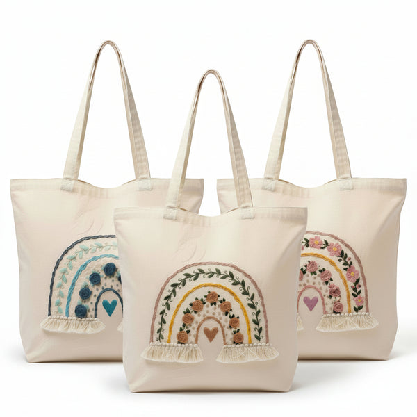 Three tote bags with embroidered designs of rainbows and hearts on a white background